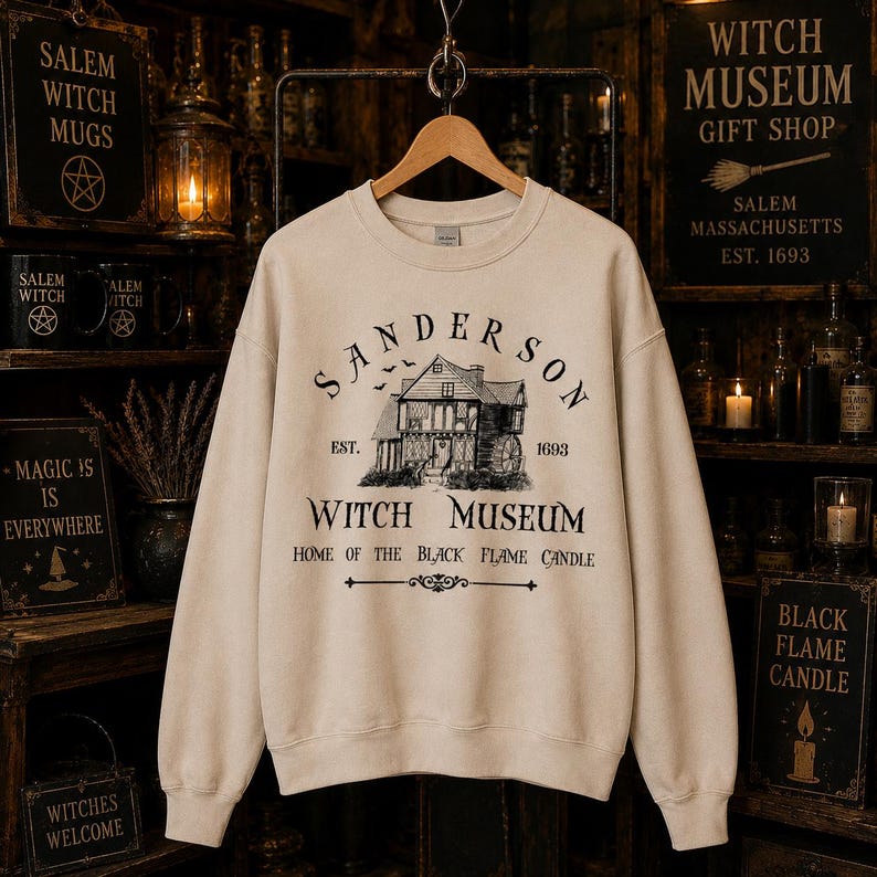 K&ouml;nnte beinhalten: Ein beigefarbenes Sweatshirt mit Rundhalsausschnitt und einem schwarzen Grafikdruck eines Hauses sowie dem Text "SANDERSON WITCH MUSEUM EST. 1693 HOME OF THE BLACK FLAME CANDLE" auf der Vorderseite. Das Design zeigt zudem kleine fliegende Flederm&auml;use.