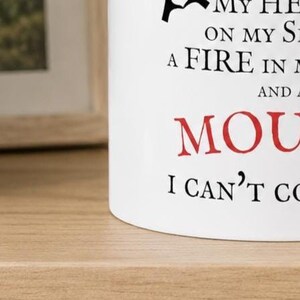 May include: A white ceramic mug featuring black and red printed text. The visible portion of the text reads: MY HE ON MY S A FIRE IN M AND A MOU I CAN'T CO. The mug has a smooth, cylindrical surface and a clean, minimalist white finish.