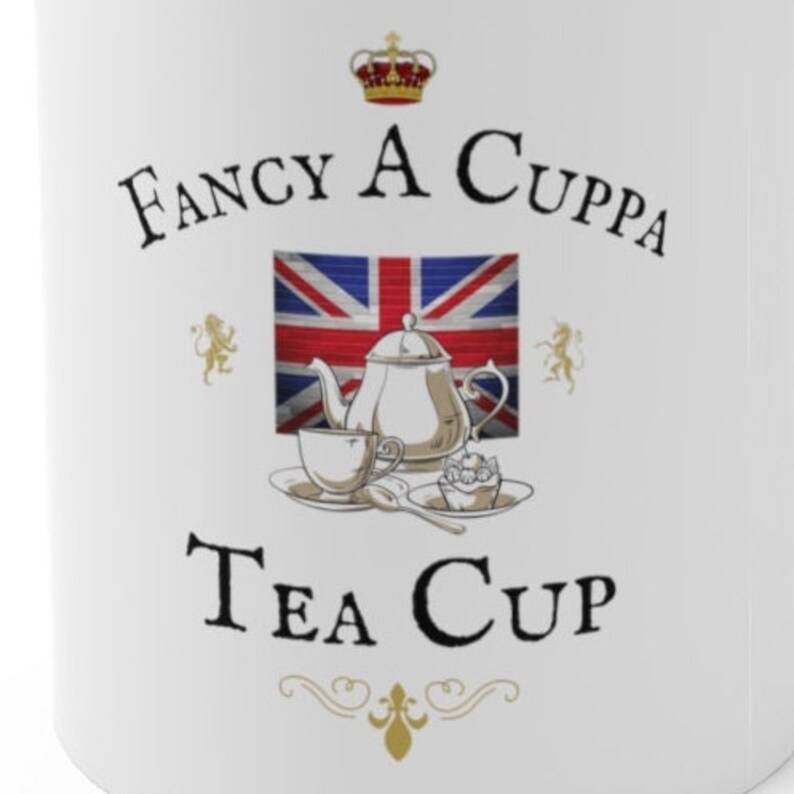 May include: A white ceramic mug featuring a graphic of a silver teapot, a teacup, a spoon, and a small cake. Behind the items is a Union Jack flag. Above the graphic is a crown and the text FANCY A CUPPA, and below is the text TEA CUP.