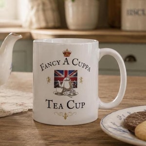 BRITISH TEA Gift Idea Teacup Funny Mug United Kingdom Gag Gift Great Britain Uk Expat Citizen Present Fancy A CUPPA Coffee Cup Union Jack Includes: A white ceramic mug featuring the text "Fancy A Cuppa Tea Cup" in black lettering. The design includes a central illustration of a teapot and teacup set against a Union Jack flag, topped with a small red crown and gold lion accents.