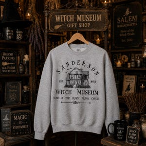 K&ouml;nnte beinhalten: Ein graumeliertes Sweatshirt mit Rundhalsausschnitt und einem schwarzen Grafikdruck eines Hauses mit dem Text Sanderson Witch Museum Est. 1693 Home of the Black Flame Candle. Das Design zeigt kleine fliegende Flederm&auml;use &uuml;ber dem Haus.