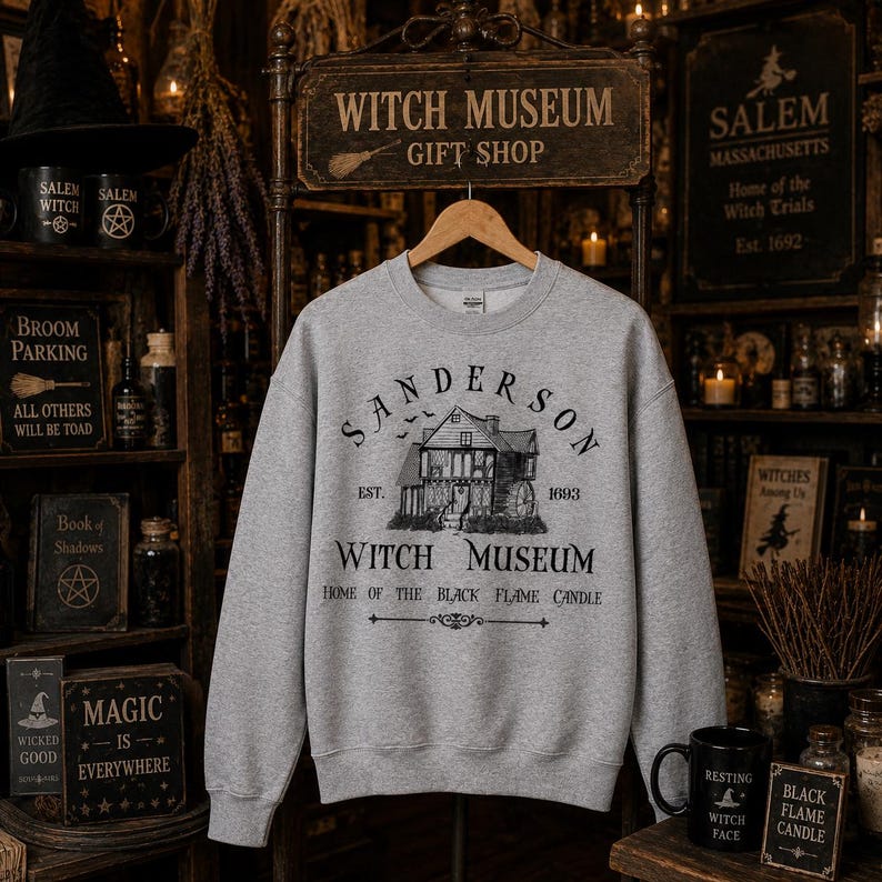 K&ouml;nnte beinhalten: Ein graumeliertes Sweatshirt mit Rundhalsausschnitt und einem schwarzen Grafikdruck eines Hauses mit dem Text Sanderson Witch Museum Est. 1693 Home of the Black Flame Candle. Das Design zeigt kleine fliegende Flederm&auml;use &uuml;ber dem Haus.