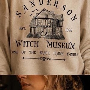 K&ouml;nnte beinhalten: Ein beigefarbenes Sweatshirt mit Rundhalsausschnitt und einem schwarzen Grafikdruck eines Hauses mit dem Text Sanderson Witch Museum, Est. 1693 und Home of the Black Flame Candle. Das Design zeigt zudem kleine fliegende Flederm&auml;use.