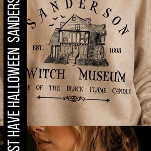 K&ouml;nnte beinhalten: Ein beigefarbenes Sweatshirt mit Rundhalsausschnitt und einem schwarzen Grafikdruck eines Hauses mit dem Text Sanderson Witch Museum, Est. 1693, und Home of the Black Flame Candle. Das Design zeigt kleine fliegende Flederm&auml;use &uuml;ber dem Haus.