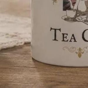 May include: A white ceramic canister featuring a black graphic illustration of a teapot, a teacup on a saucer, and a spoon. The text "TEA C" is printed in a black serif font on the front, accompanied by a decorative gold-colored flourish design.