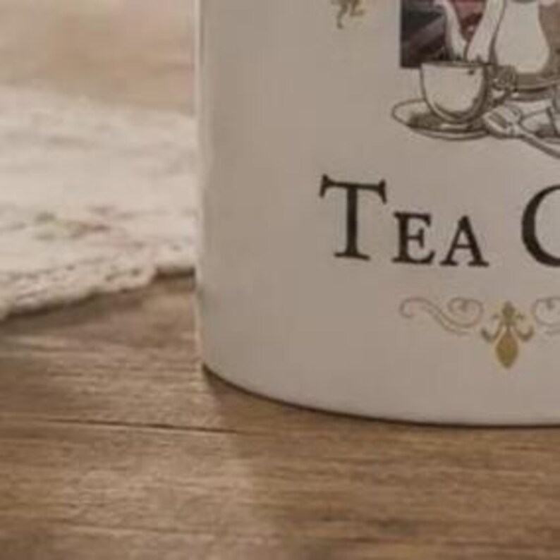 May include: A white ceramic canister featuring a black graphic illustration of a teapot, a teacup on a saucer, and a spoon. The text "TEA C" is printed in a black serif font on the front, accompanied by a decorative gold-colored flourish design.