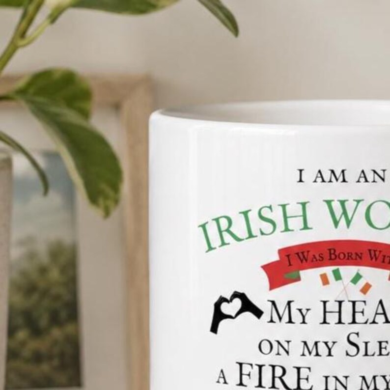 May include: A white ceramic mug featuring black and green text that reads I AM AN IRISH WOMAN I WAS BORN WITH MY HEART ON MY SLEEVE A FIRE IN MY SOUL. The design includes a red banner, two small crossed Irish flags, and a black heart-shaped hand icon.