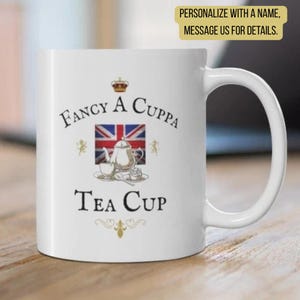 May include: A white ceramic mug featuring a graphic of a silver teapot on a saucer in front of a Union Jack flag. The text reads "FANCY A CUPPA TEA CUP" with a small crown icon above and decorative flourishes below the design on the mug surface.