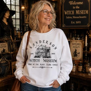 K&ouml;nnte beinhalten: Ein wei&szlig;es lang&auml;rmeliges Sweatshirt mit Rundhalsausschnitt und einem schwarzen Grafikdruck eines Hauses mit dem Text Sanderson Witch Museum Est. 1693 Home of the Black Flame Candle. Das Design enth&auml;lt eine kleine Abbildung fliegender Flederm&auml;use.