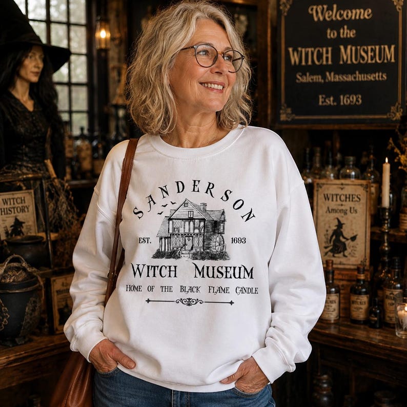 K&ouml;nnte beinhalten: Ein wei&szlig;es lang&auml;rmeliges Sweatshirt mit Rundhalsausschnitt und einem schwarzen Grafikdruck eines Hauses mit dem Text Sanderson Witch Museum Est. 1693 Home of the Black Flame Candle. Das Design enth&auml;lt eine kleine Abbildung fliegender Flederm&auml;use.