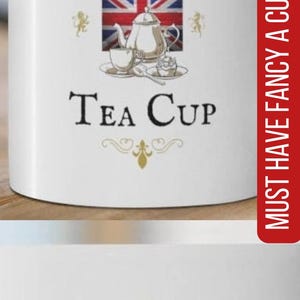 May include: A white ceramic mug featuring a graphic of a Union Jack flag, a silver teapot, a teacup, and a cupcake. The text reads "Fancy A Cuppa" above the flag and "Tea Cup" below, with a small crown icon at the top and gold flourishes.