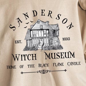 K&ouml;nnte beinhalten: Ein beiges T-Shirt mit einer schwarzen Tuschezeichnung eines Hauses mit Wasserrad. Der Text lautet: SANDERSON EST. 1693 WITCH MUSEUM HOME OF THE BLACK FLAME CANDLE. &Uuml;ber dem Dach des Hauses sind drei kleine Flederm&auml;use abgebildet.