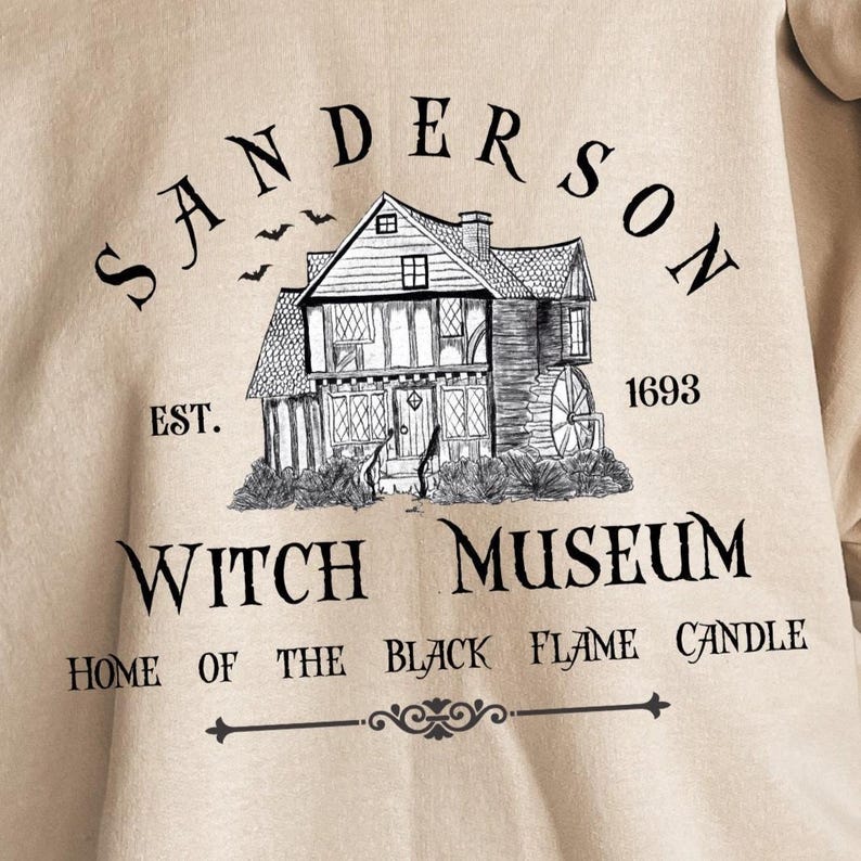 K&ouml;nnte beinhalten: Ein beiges T-Shirt mit einer schwarzen Tuschezeichnung eines Hauses mit Wasserrad. Der Text lautet: SANDERSON EST. 1693 WITCH MUSEUM HOME OF THE BLACK FLAME CANDLE. &Uuml;ber dem Dach des Hauses sind drei kleine Flederm&auml;use abgebildet.