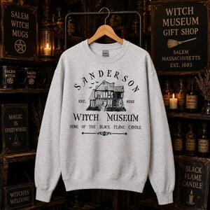 K&ouml;nnte beinhalten: Ein hellgraues Sweatshirt mit Rundhalsausschnitt und einem schwarzen Grafikdruck eines Hauses mit dem Text Sanderson Witch Museum Est. 1693 Home of the Black Flame Candle. Das Design enth&auml;lt kleine Fledermaussilhouetten &uuml;ber dem Haus.