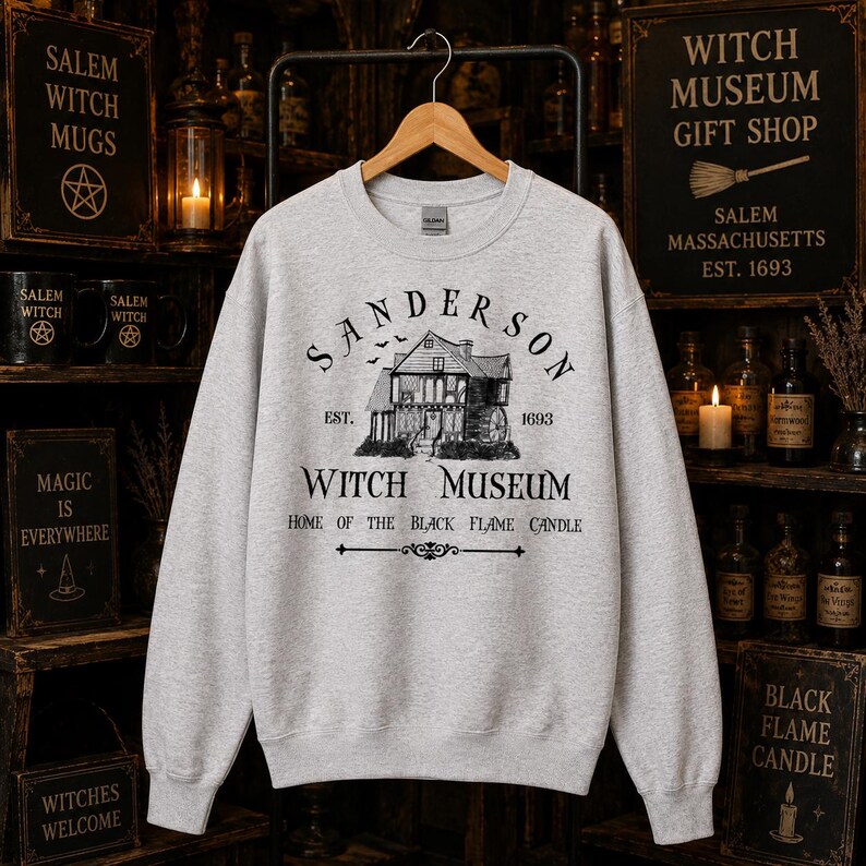 K&ouml;nnte beinhalten: Ein hellgraues Sweatshirt mit Rundhalsausschnitt und einem schwarzen Grafikdruck eines Hauses mit dem Text Sanderson Witch Museum Est. 1693 Home of the Black Flame Candle. Das Design enth&auml;lt kleine Fledermaussilhouetten &uuml;ber dem Haus.