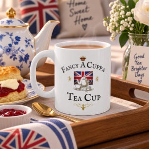 May include: A white ceramic mug filled with tea, featuring the text "Fancy A Cuppa Tea Cup" in black lettering, a small crown icon, a Union Jack flag graphic, and a detailed illustration of a teapot, teacup, and sugar bowl in the center.
