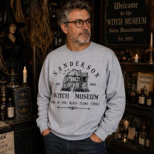 K&ouml;nnte beinhalten: Ein hellgraues Sweatshirt mit Rundhalsausschnitt und einem schwarzen Grafikdruck eines Hauses mit dem Text Sanderson Witch Museum Est. 1693 Home of the Black Flame Candle. Das Design enth&auml;lt eine kleine Abbildung eines Hauses mit Wasserrad.
