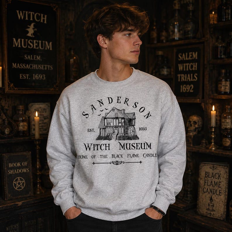 K&ouml;nnte beinhalten: Ein hellgraues Sweatshirt mit Rundhalsausschnitt und einem schwarzen Grafikdruck eines Hauses mit dem Text Sanderson Witch Museum, Est. 1693, und Home of the Black Flame Candle. Das Design enth&auml;lt eine kleine Illustration eines Hauses und fliegender Flederm&auml;use.