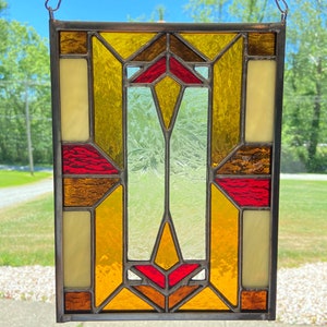K&ouml;nnte beinhalten: Ein Buntglasfenster mit einem geometrischen Muster aus gelben, orangefarbenen und roten Glasscheiben. Das Fenster ist in Metall gefasst und h&auml;ngt an einer Kette.