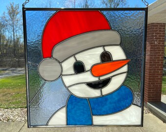 May include: A stained glass window depicting a snowman wearing a red and white hat and a blue scarf. The snowman has a carrot nose and black eyes.