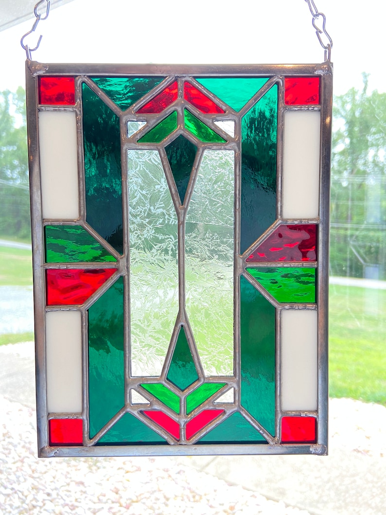 K&ouml;nnte beinhalten: Ein Buntglasfenster mit einem geometrischen Muster aus roten, gr&uuml;nen und wei&szlig;en Glasscheiben. Das Fenster ist in silberfarbenem Metall gefasst und h&auml;ngt an einer Kette.