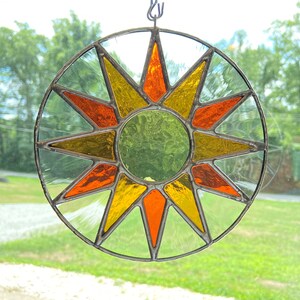 May include: A stained glass sun catcher featuring a 12-pointed star design. The star is made from clear, orange, yellow, and green glass.