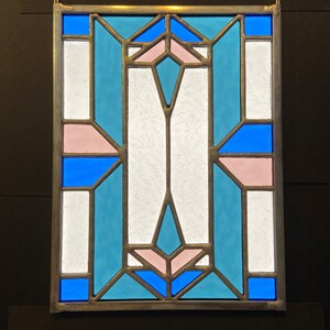 K&ouml;nnte beinhalten: Ein Buntglasfenster mit einem geometrischen Muster aus blauen, rosa und wei&szlig;en Glasscheiben.