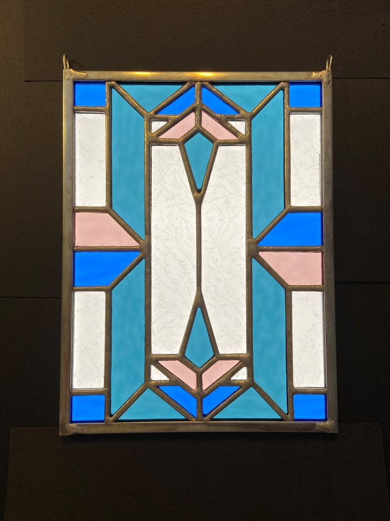 K&ouml;nnte beinhalten: Ein Buntglasfenster mit einem geometrischen Muster aus blauen, rosa und wei&szlig;en Glasscheiben.