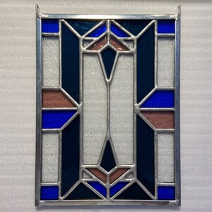 K&ouml;nnte beinhalten: Ein Bleiglasfenster mit einem geometrischen Muster aus blauen, dunkelblauen und braunen Glasscheiben. Das Fenster ist in silberfarbenem Metall gerahmt.