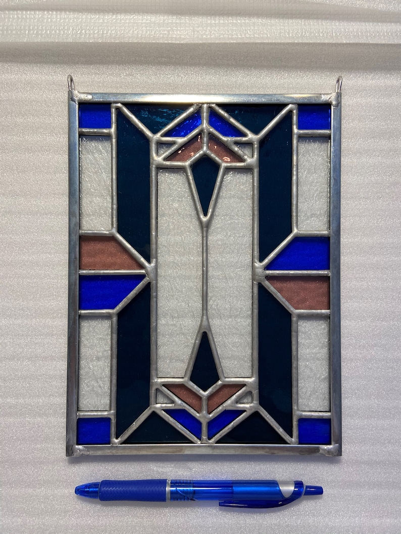 K&ouml;nnte beinhalten: Ein Bleiglasfenster mit einem geometrischen Muster aus blauen, dunkelblauen und braunen Glasscheiben. Das Fenster ist in silberfarbenem Metall gerahmt.