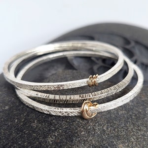 May include: Three silver bangles with a textured finish, stacked on top of each other. The top bangle has a small gold knot accent, and the middle bangle has a gold wire accent.