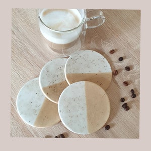 May include: Four round, speckled, beige and white coasters with a coffee cup on top. The coasters are made of a speckled material and have a light brown and white color scheme.