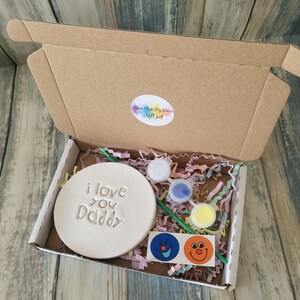 A craft kit for making a personalised gift. The kit includes a white ceramic round plaque with the words "i love you Daddy" on it, paint, a paintbrush, and two smiley face stickers.