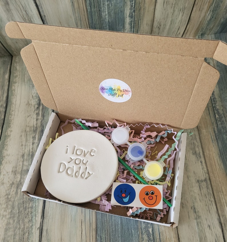 A craft kit for making a personalised gift. The kit includes a white ceramic round plaque with the words "i love you Daddy" on it, paint, a paintbrush, and two smiley face stickers.