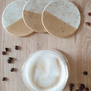 May include: Set of four round ceramic coasters with a white and beige speckled design. The coasters are arranged in a stack with the top coaster showing a white top half and a beige bottom half.