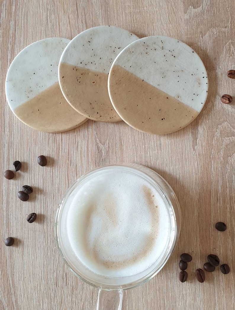 May include: Set of four round ceramic coasters with a white and beige speckled design. The coasters are arranged in a stack with the top coaster showing a white top half and a beige bottom half.