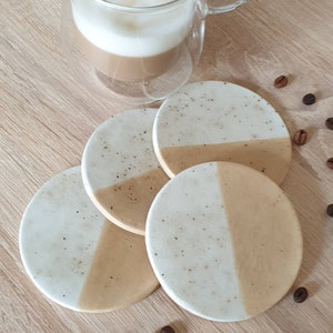 May include: Four round coasters with a white top and a beige bottom. The coasters have a speckled pattern and are arranged in a circle.