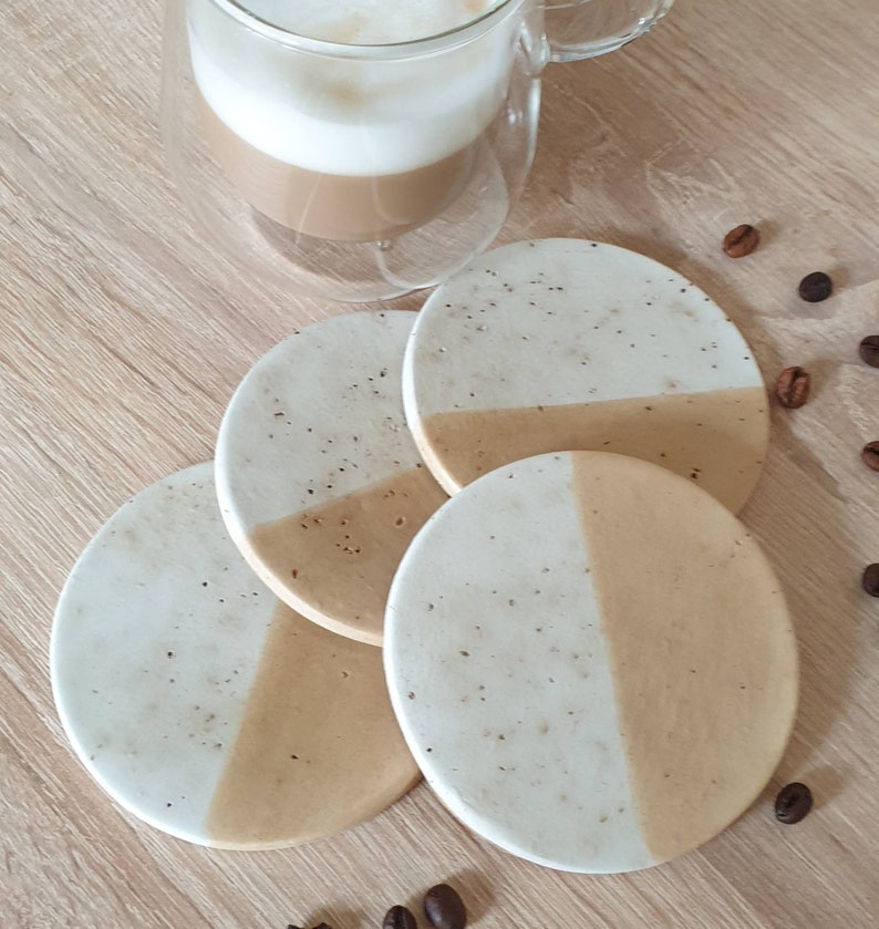 May include: Four round coasters with a white top and a beige bottom. The coasters have a speckled pattern and are arranged in a circle.