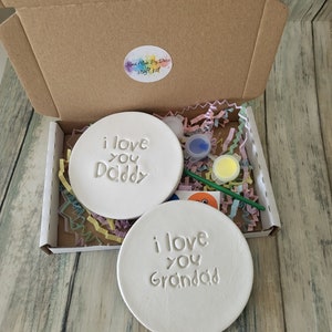 Puede incluir: Un kit de manualidades con dos posavasos de cer&aacute;mica blanca, uno con el texto "Te quiero pap&aacute;" y el otro con el texto "Te quiero abuelo". El kit incluye pintura, un pincel y una pegatina.