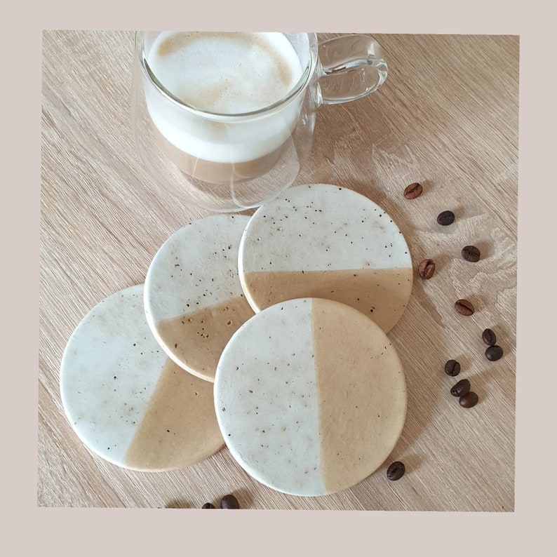 May include: Four round, speckled, beige and white coasters with a coffee cup on top. The coasters are made of a speckled material and have a light brown and white color scheme.