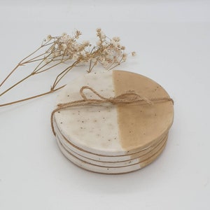 May include: Set of four ceramic coasters with a white and beige speckled glaze. The coasters are round and have a half-and-half design, with one half white and the other half beige.