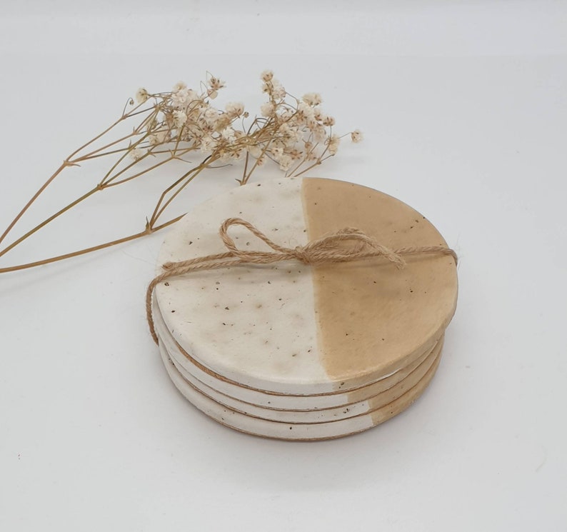 May include: Set of four ceramic coasters with a white and beige speckled glaze. The coasters are round and have a half-and-half design, with one half white and the other half beige.