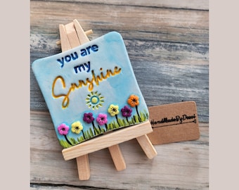 May include: A small blue ceramic sign with the text "You are my Sunshine" in yellow lettering. The sign is decorated with a yellow sun and colorful flowers and is displayed on a small wooden easel. The sign has a handmade label.