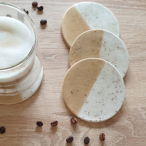 May include: Set of four round coasters with a white and beige speckled design. Each coaster is divided in half, with one half white and the other half beige.