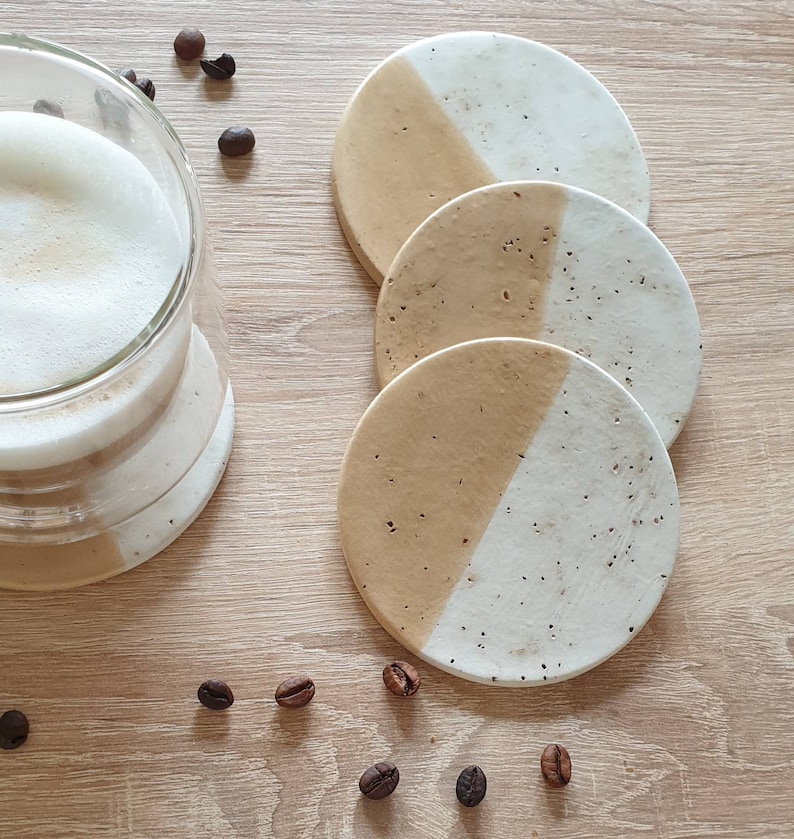 May include: Set of four round coasters with a white and beige speckled design. Each coaster is divided in half, with one half white and the other half beige.