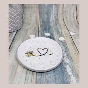 May include: White ceramic ring dish with a black and yellow bee design and a black heart drawn on the surface.