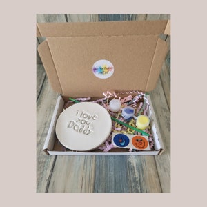 May include: A craft kit for making a personalised gift. The kit includes a white ceramic disc with the words "I love you Daddy" engraved on it, two small paint pots, a paintbrush, and two smiley face stickers. The kit is packaged in a brown cardboard box with a sticker that says "Paint Your Own Dad Craft Kit".