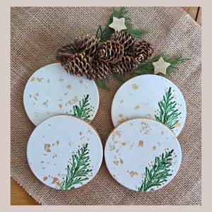 May include: Four white ceramic coasters with gold flecks and a green painted evergreen branch design. The coasters are round and have a gold rim. The image also includes pine cones and greenery, suggesting a festive theme.