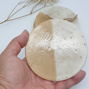 May include: Set of two round ceramic coasters with a white and beige glaze. The coasters are half white and half beige, with a rough texture.