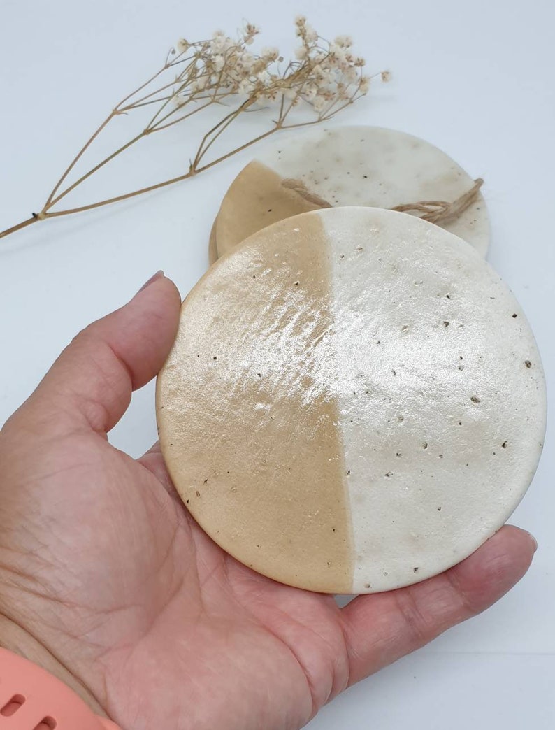 May include: Set of two round ceramic coasters with a white and beige glaze. The coasters are half white and half beige, with a rough texture.